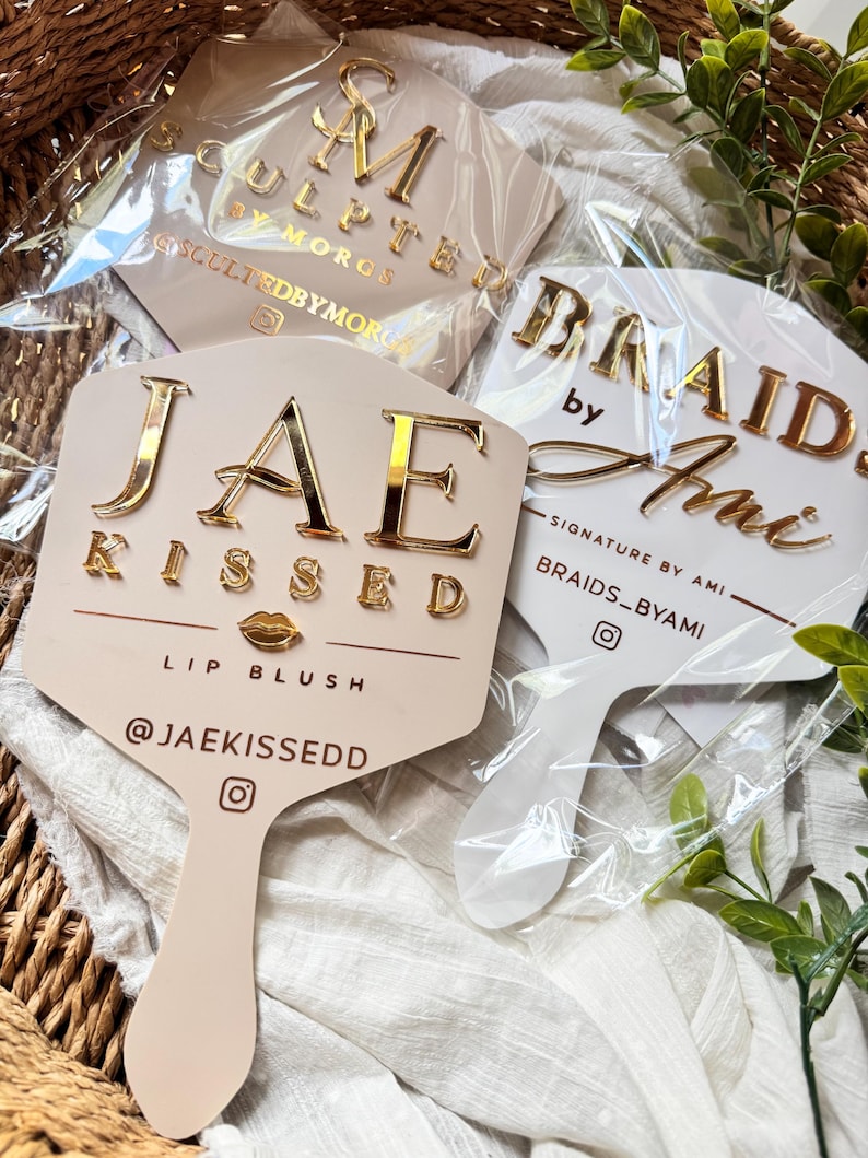May include: Three handheld hexagonal mirrors with custom gold-toned lettering. One reads JAE KISSED LIP BLUSH @JAEKISSEDD, another BRAIDS BY AMI SIGNATURE BY AMI BRAIDS_BYAMI, and the third SCULPTED BY MORGS SCULPTEDBYMORGS, all encased in clear plastic.