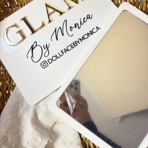 K&ouml;nnte beinhalten: Ein rechteckiges wei&szlig;es Schild mit dem goldfarbenen Schriftzug GLAM By Monica und dem schwarzen Text DOLLFACEBYMONICA samt Instagram-Symbol. An dem Schild ist ein rechteckiger Spiegel mit einer teilweise abgezogenen Schutzfolie befestigt.