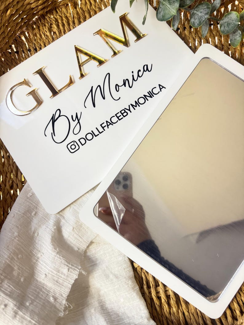 K&ouml;nnte beinhalten: Ein rechteckiges wei&szlig;es Schild mit dem goldfarbenen Schriftzug GLAM By Monica und dem schwarzen Text DOLLFACEBYMONICA samt Instagram-Symbol. An dem Schild ist ein rechteckiger Spiegel mit einer teilweise abgezogenen Schutzfolie befestigt.