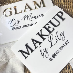 K&ouml;nnte beinhalten: Zwei rechteckige wei&szlig;e Acrylschilder mit erhabener Beschriftung. Ein Schild zeigt den goldenen Schriftzug GLAM By Monica und den Instagram-Namen DOLLFACEBYM. Das zweite Schild zeigt schwarzen Text MAKEUP by Lily und @HMUBYLILY.