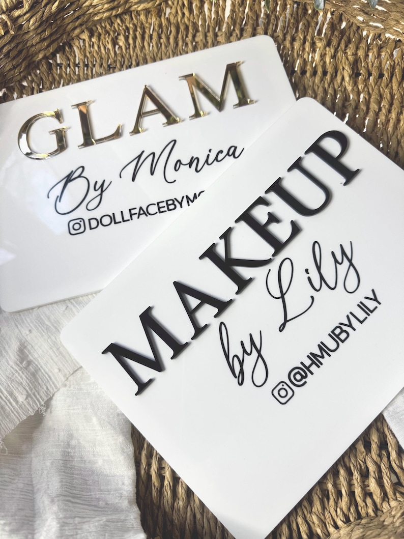 K&ouml;nnte beinhalten: Zwei rechteckige wei&szlig;e Acrylschilder mit erhabener Beschriftung. Ein Schild zeigt den goldenen Schriftzug GLAM By Monica und den Instagram-Namen DOLLFACEBYM. Das zweite Schild zeigt schwarzen Text MAKEUP by Lily und @HMUBYLILY.