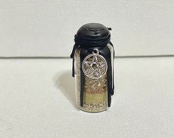 May include: A small glass jar with a black lid and a black ribbon tied around the top. The jar is filled with dried herbs and spices. A silver pentagram charm hangs from the ribbon.