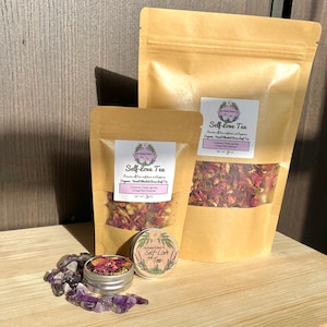 May include: Two brown paper bags with a label that reads "Self-love Tea" and a smaller tin with the same label. The bags contain loose leaf tea with dried rose petals and other herbs. The tin contains a small amount of the tea. There are also amethyst crystals scattered on a wooden surface.