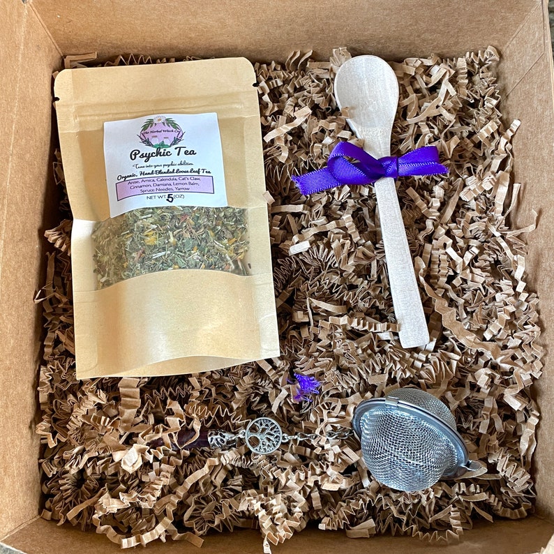 May include: A brown cardboard box filled with brown packing paper. The box contains a small brown pouch of tea labeled "Psychic Tea", a wooden spoon tied with a purple ribbon, a metal tea infuser, and a silver charm with a tree design.
