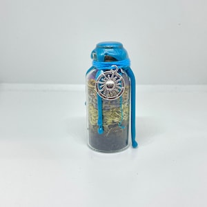 May include: A small glass bottle with a blue wax seal and a silver sun charm. The bottle is filled with dried herbs and flowers.