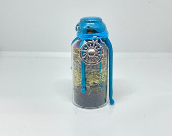 Puede incluir: Una pequeña botella de vidrio con un sello de cera azul y un amuleto de sol plateado. La botella está llena de hierbas y flores secas.