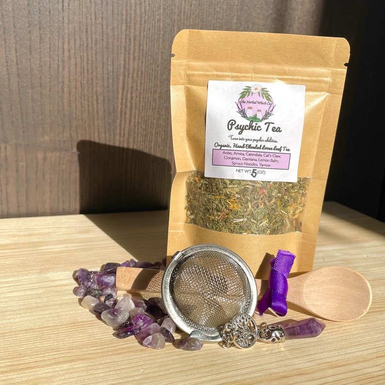 May include: A brown paper bag with a label that reads "Psychic Tea" and lists the ingredients: Anise, Amica, Calendula, Cat's Claw, Cinnamon, Damiana, Lemon Balm, Spruce Needles, Yarrow. The bag contains a blend of dried herbs. The bag is sitting on a wooden surface with a purple amethyst crystal necklace, a wooden spoon, and a metal tea infuser.