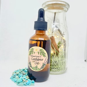 May include: A brown glass bottle with a black dropper, labeled "Confidence Oil" with floral illustrations. A clear glass jar with fairy lights, a cinnamon stick, and greenery is behind the bottle. Turquoise stones are in front.
