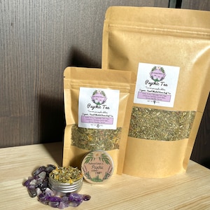 May include: Two brown paper bags filled with loose leaf tea, labeled "Psychic Tea" with a list of ingredients. A smaller tin container with the same label is in front of the bags. The bags and tin are on a wooden surface with purple amethyst stones scattered around.