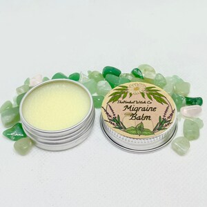 May include: A small silver tin with a label that reads "The Herbal Witch Co. Migraine Balm". The label features a floral design with a white daisy and green leaves. The tin is open and the balm is visible inside.