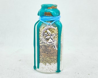 May include: A small glass bottle filled with herbs and crystals, sealed with a teal wax and a cork stopper. A silver charm of a fairy on a crescent moon hangs from a blue string around the neck. The bottle contains layers of white, green, and brown materials.