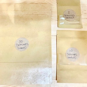 May include: Three brown paper bags with labels indicating different serving sizes. The largest bag is labeled "30 Servings (Large)", the medium bag is labeled "10 Servings (Medium)", and the smallest bag is labeled "2 Serving Sampler".