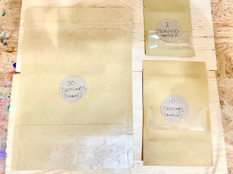 May include: Three brown paper bags with labels indicating different serving sizes. The largest bag is labeled "30 Servings (Large)", the medium bag is labeled "10 Servings (Medium)", and the smallest bag is labeled "2 Serving Sampler".