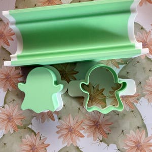 May include: A light green and white cylindrical rolling pin guide and two ghost-shaped cookie cutters. The rolling pin guide is a long, curved piece, while the two ghost cutters feature a solid light green top and a white base frame.