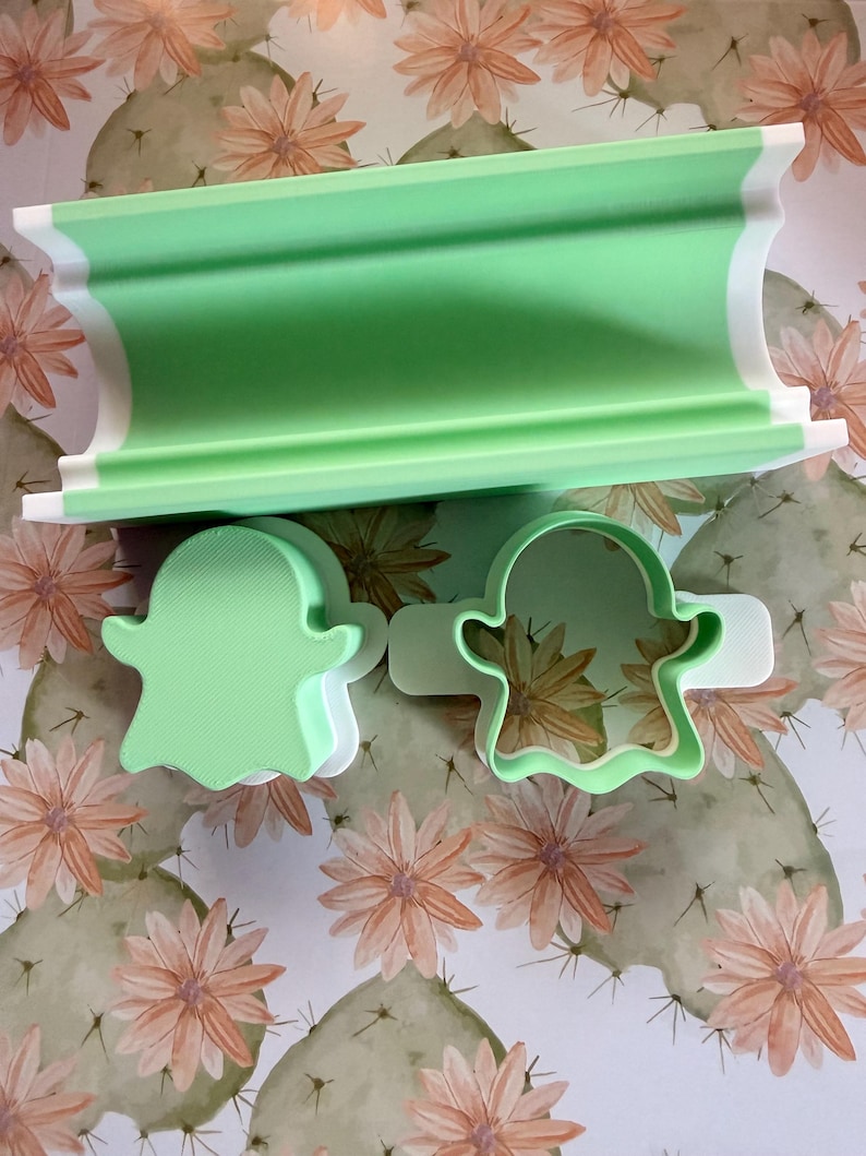 May include: A light green and white cylindrical rolling pin guide and two ghost-shaped cookie cutters. The rolling pin guide is a long, curved piece, while the two ghost cutters feature a solid light green top and a white base frame.