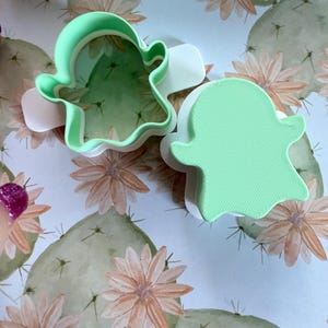 May include: Two ghost-shaped cookie cutters made of light green and white plastic. One cutter is open, showing the interior walls, while the other is closed with a solid top surface featuring a textured, linear pattern across the ghost shape.