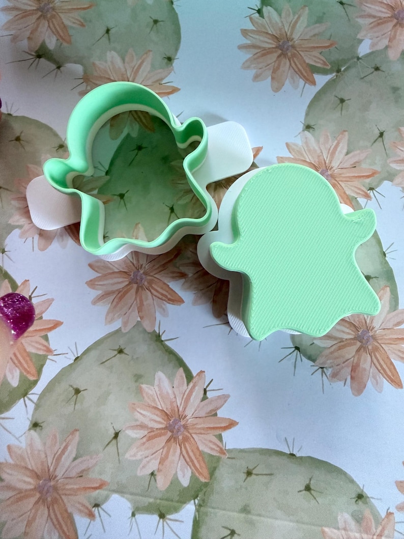 May include: Two ghost-shaped cookie cutters made of light green and white plastic. One cutter is open, showing the interior walls, while the other is closed with a solid top surface featuring a textured, linear pattern across the ghost shape.