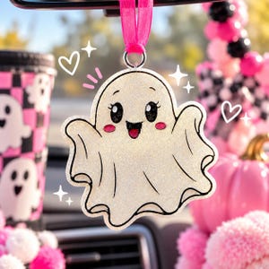 May include: A glittery white ghost ornament with a cheerful expression, black eyes, and pink cheeks, suspended by a bright pink ribbon. The ghost features a cartoon-style design with black outlines and is accented by white hand-drawn heart and star icons.