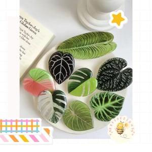 May include: A collection of leaf-shaped decorative items in various shades of green, white, and pink, arranged on a white plate. The leaves have detailed veining and glossy surfaces, resembling different plant species. A book and a bee sticker are also visible.