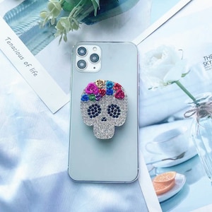 May include: A white phone case with a rhinestone skull phone grip. The skull is decorated with colorful rhinestones and has a floral design on the top of the head.