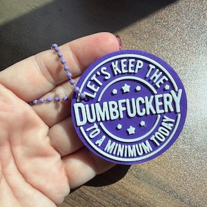 May include: A circular purple plastic tag with white raised lettering that reads "LET'S KEEP THE DUMBFUCKERY TO A MINIMUM TODAY" with small star accents. The tag is attached to a light purple ball chain, featuring a textured 3D printed finish.