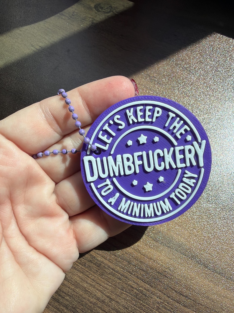 May include: A circular purple plastic tag with white raised lettering that reads "LET'S KEEP THE DUMBFUCKERY TO A MINIMUM TODAY" with small star accents. The tag is attached to a light purple ball chain, featuring a textured 3D printed finish.