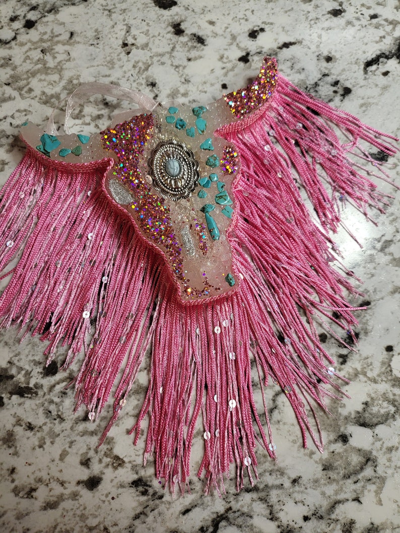 May include: A decorative bull skull ornament with pink fringe and turquoise accents. The skull is embellished with glitter and a silver-toned centerpiece. A clear ribbon is attached for hanging.