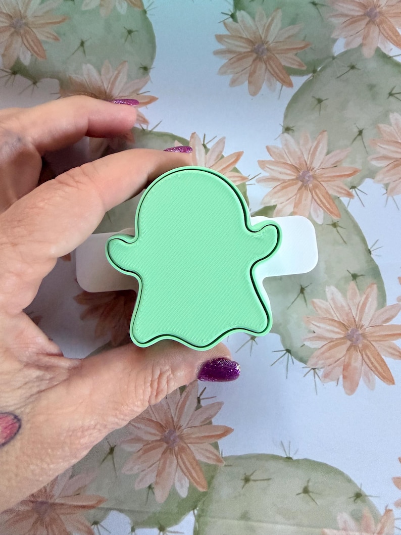 May include: A light green ghost-shaped cookie cutter with a smooth, flat top surface and a white base. The cutter features a classic ghost silhouette with rounded edges and a defined, raised border, designed for cutting dough into a ghost shape.