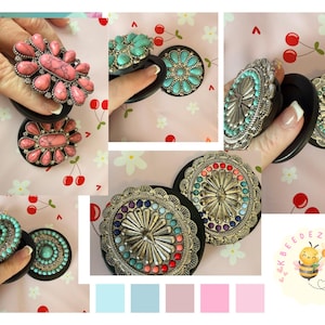 May include: A collection of decorative phone grips with various designs. The grips feature silver-toned metalwork and colorful stone accents in shades of pink, turquoise, and other colors. The background is a light pink with a cherry pattern.