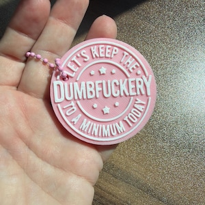 May include: A circular pink keychain with raised white lettering that reads "LET'S KEEP THE DUMBFUCKERY TO A MINIMUM TODAY" with small star accents. The keychain features a pink ball chain attachment and has a textured, layered appearance.