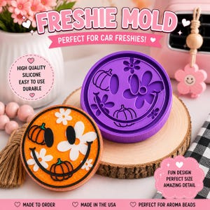 May include: A round purple silicone mold featuring a smiley face design with pumpkins and flowers, alongside a finished orange circular air freshener with a matching smiley face, pumpkin, and flower pattern, with text reading FRESHIE MOLD.