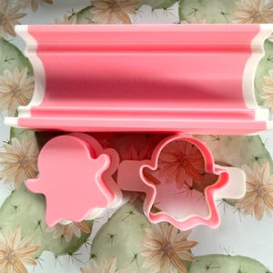 May include: A pink and white rectangular press tool alongside two ghost-shaped cookie cutters. One cutter is a solid pink ghost shape, while the other is a pink and white outline of a ghost with a handle on the side for easy gripping and use.
