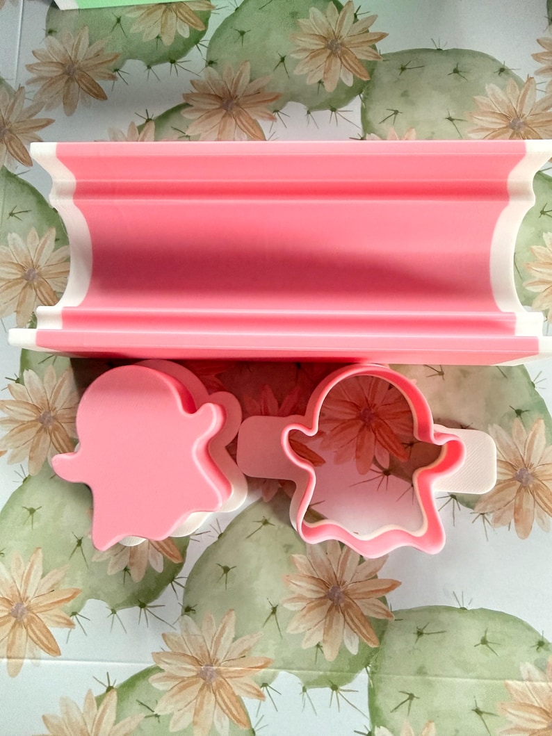 May include: A pink and white rectangular press tool alongside two ghost-shaped cookie cutters. One cutter is a solid pink ghost shape, while the other is a pink and white outline of a ghost with a handle on the side for easy gripping and use.