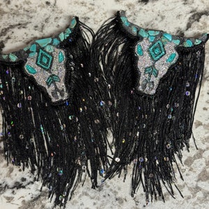 May include: A pair of black tassel earrings with turquoise and silver skull charms. The skulls are embellished with glitter and rhinestones.