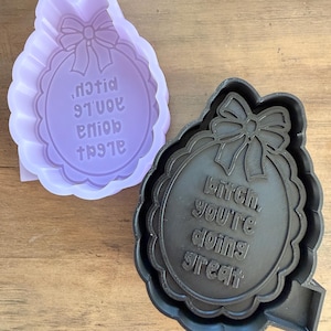 May include: Two cookie cutters, one lavender and one black, with a scalloped edge and bow design. The text "Bitch, you're doing great" is embossed on the black cutter. The lavender cutter has the same design, but the text is reversed.