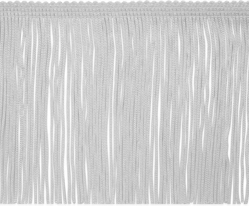 May include: Close-up of a white fringe trim. The trim features a woven top edge with a scalloped design and long, vertical strands of fringe. The fringe is a soft, light color, suitable for various craft and design projects.