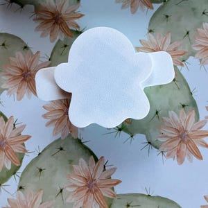 May include: A white, ghost-shaped adhesive bandage with a smooth, matte texture. The bandage features a central rounded body with two horizontal side tabs extending outward, designed for covering small wounds or skin abrasions on the body.