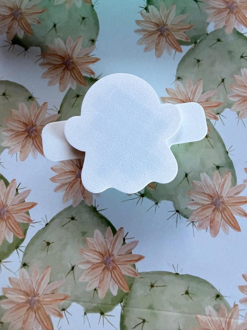 May include: A white, ghost-shaped adhesive bandage with a smooth, matte texture. The bandage features a central rounded body with two horizontal side tabs extending outward, designed for covering small wounds or skin abrasions on the body.