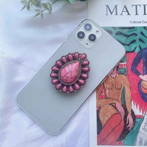 May include: A pink and silver phone ring holder with a teardrop-shaped stone. The stone has a marbled pattern and is surrounded by smaller pink stones.