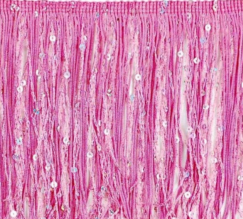 May include: Close-up of a pink fringe trim with a dense, textured appearance. The trim is made of long, vertical strands of pink material, interspersed with small, shimmering sequins. The fringe is likely used for embellishing clothing or accessories.