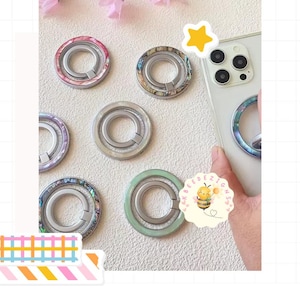 May include: A collection of phone ring holders in various colors and designs. The rings feature iridescent and solid color finishes, with a circular design and a metal ring for grip. One is attached to a phone.