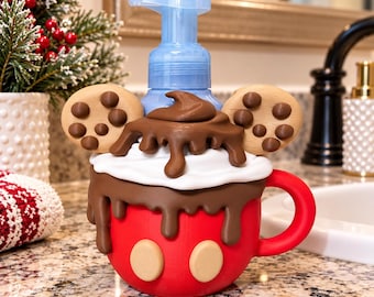 May include: A red soap dispenser shaped like a mug with a handle, topped with white whipped cream, brown chocolate drizzle, and two round chocolate chip cookies. The base features two beige circles, and a light blue pump mechanism is attached to the top.