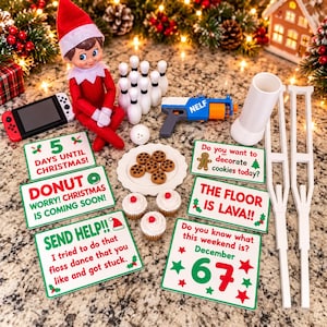 May include: A Christmas elf doll sits among miniature toys including a bowling set, a toy blaster, a handheld gaming console, three cupcakes, a plate of cookies, two white crutches, and several signs with holiday-themed text and graphics.