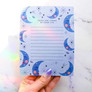 Peut inclure: Bloc-notes bleu et blanc avec un motif de lune et d'étoiles. Le bloc-notes a du papier ligné et le texte "Trust the timing of your life" en haut.