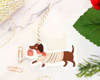 May include: A white gift tag with a brown and white dachshund wearing a pink and white sweater and carrying a red gift box. The tag has a hole at the top for string.