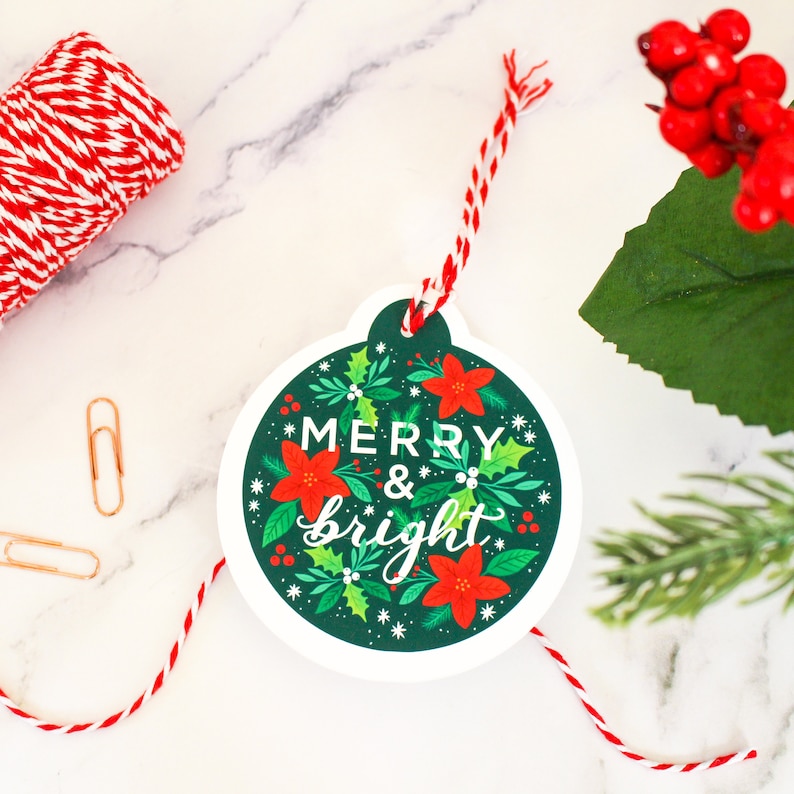 May include: A white gift tag shaped like a Christmas ornament with a green background and red poinsettia flowers. The tag has the text "Merry & bright" in white lettering.