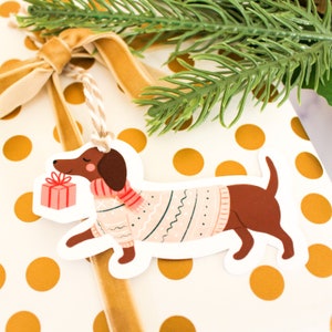May include: A white gift tag shaped like a dachshund wearing a pink sweater and a red and white scarf. The dachshund is holding a small pink gift box. The tag has a hole at the top for string.