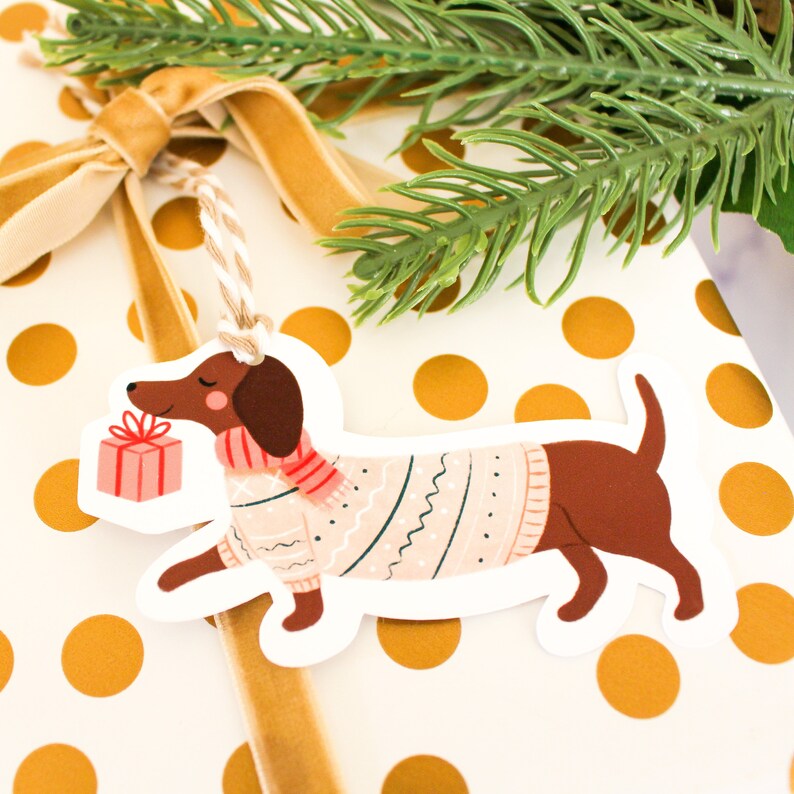 May include: A white gift tag shaped like a dachshund wearing a pink sweater and a red and white scarf. The dachshund is holding a small pink gift box. The tag has a hole at the top for string.