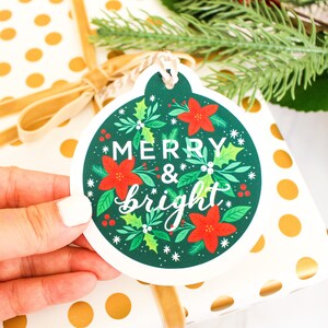 May include: A green and white Christmas gift tag shaped like an ornament with red poinsettia flowers and green holly leaves. The tag says "Merry & bright" in white lettering.