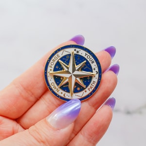 May include: Gold compass pin with a blue and white enamel design. The compass features a star with a white center and gold points. The text around the compass reads "Forge Your Own Path".